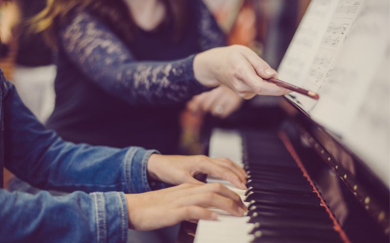How Much Do Music Lessons Cost? A piano teacher working with her student on a piece of music at the piano
