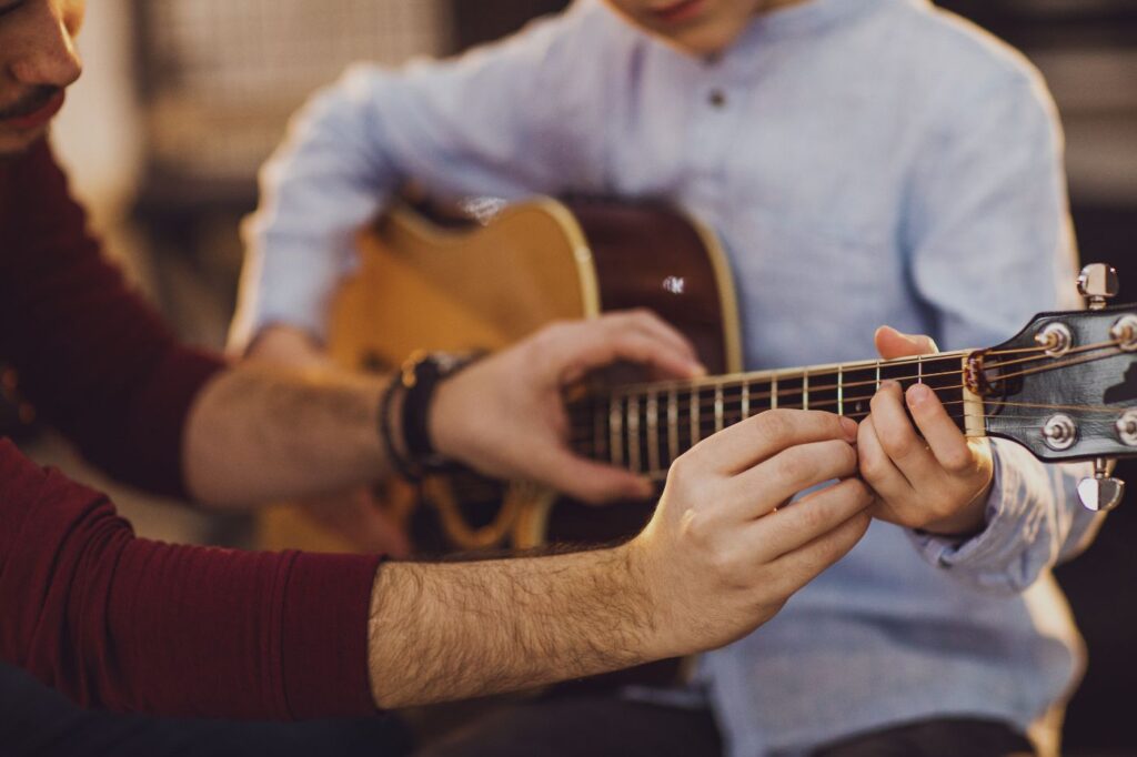 A young man taking guitar lessons from a guitar teacher that is helping with the first step of placing the student's hands correctly on the guitar neck.