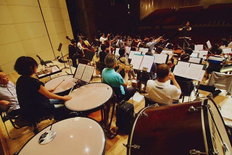 Evolution of musical form and instruments - an orchestra rehearsing on stage, with the percussionist on the forefront and the rest of the players and the conductor visible in the background.