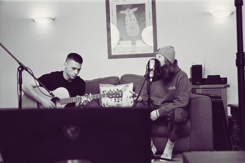 Independent artists sitting on a couch in their living room with microphones set up to record their performance.